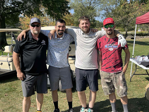 Four golfers smiling together in front of a golf cart at the annual company outing.
