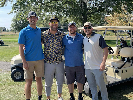 Four golfers smiling together in front of a golf cart at the annual company outing.