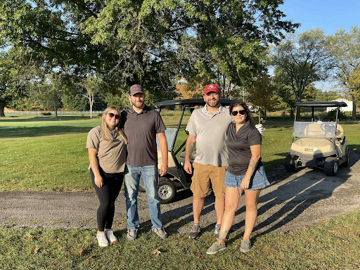 Four golfers smiling together in front of a golf cart at the annual company outing.