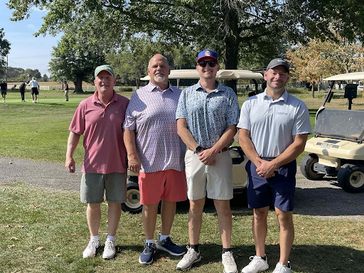 Four golfers smiling together in front of a golf cart at the annual company outing.