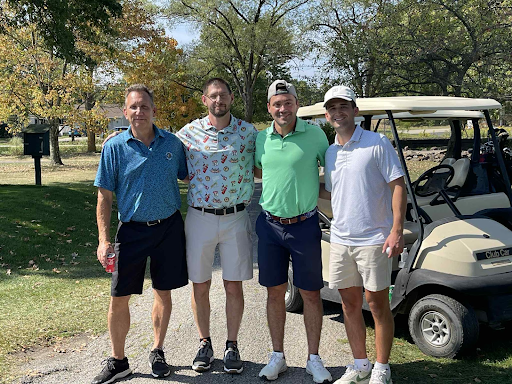 Four golfers smiling together in front of a golf cart at the annual company outing.