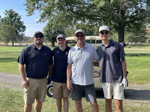 Four golfers smiling together in front of a golf cart at the annual company outing.